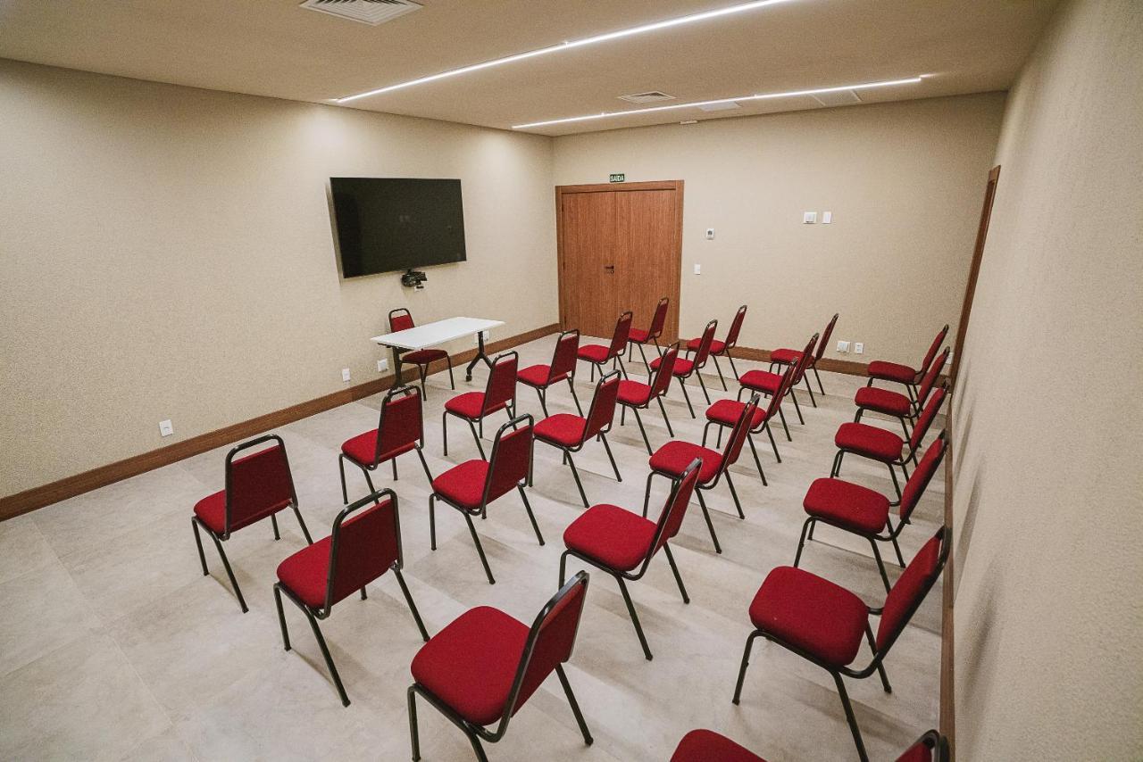 Buona Vitta Gramado Resort & Spa By Gramado Parks Екстер'єр фото The photo shows a conference or meeting room with a simple and modern design. There are several rows of red chairs arranged in a semi-circle facing a white table at the front. On the wall opposite the chairs, there is a flat-screen television mounted