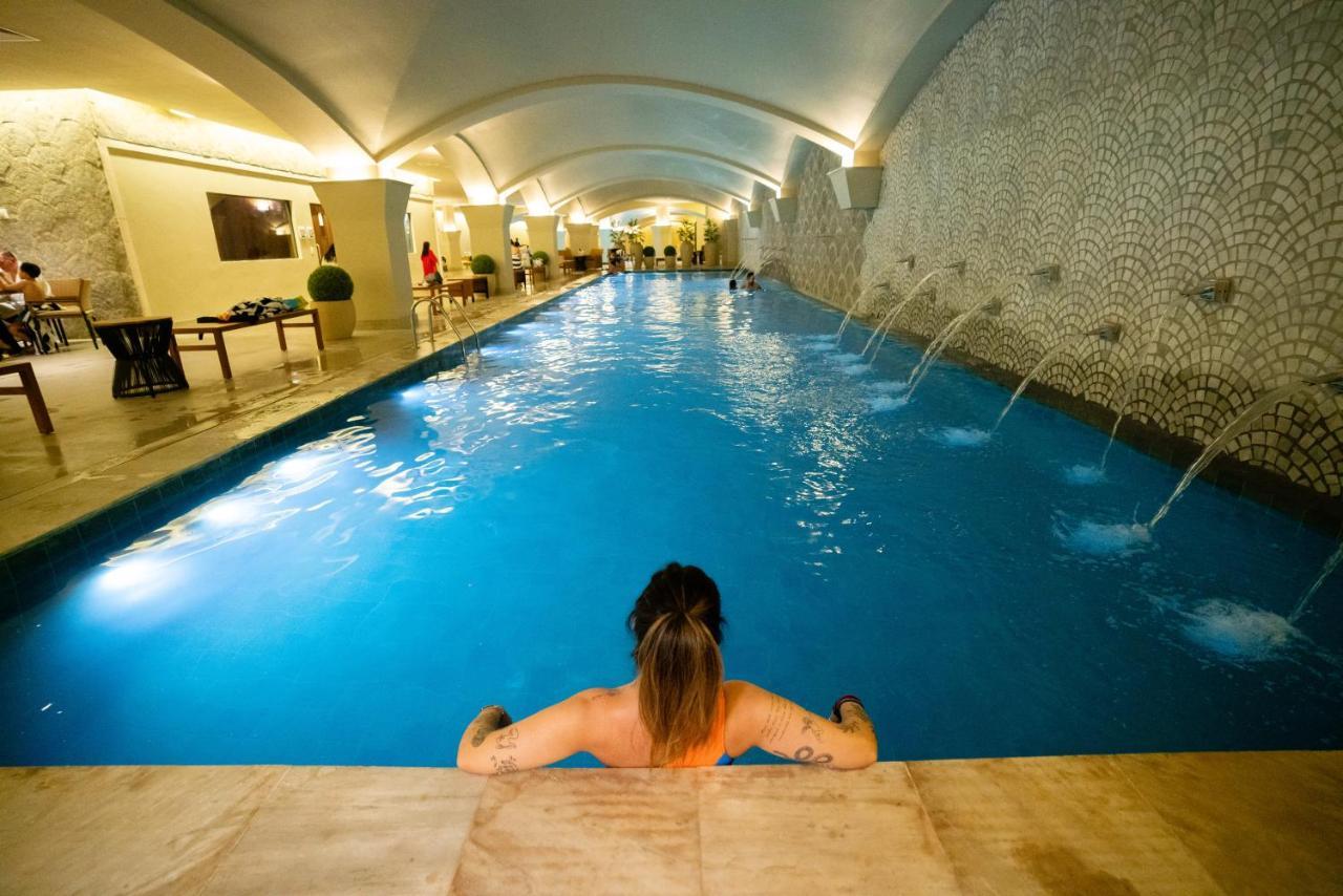 Buona Vitta Gramado Resort & Spa By Gramado Parks Екстер'єр фото The photo shows an indoor swimming pool with a long, narrow design. The water is a clear blue, and there are features such as fountains along one side. In the foreground, a person with long hair is sitting on the edge of the pool, looking out over th