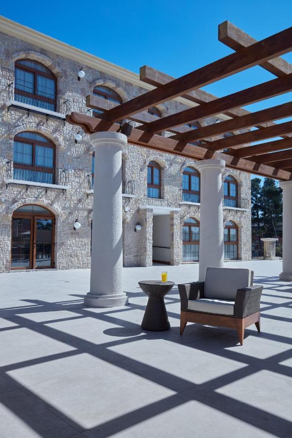Buona Vitta Gramado Resort & Spa By Gramado Parks Екстер'єр фото The photo shows a well-designed outdoor space with a stone wall building in the background. There are several tall columns that support a wooden pergola overhead, creating a shaded area. In this space, there is a stylish chair and a small table besid