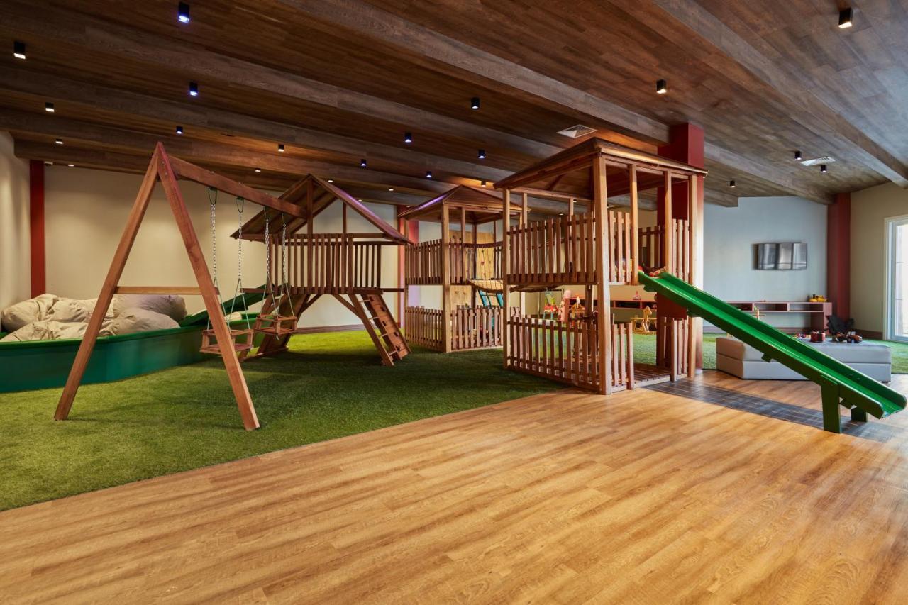 Buona Vitta Gramado Resort & Spa By Gramado Parks Екстер'єр фото The photo shows an indoor play area designed for children. It features a wooden play structure that includes multiple levels, slides, and a swing set. The floor is covered with wooden planks, and there is a green carpet-like area that adds a soft tou