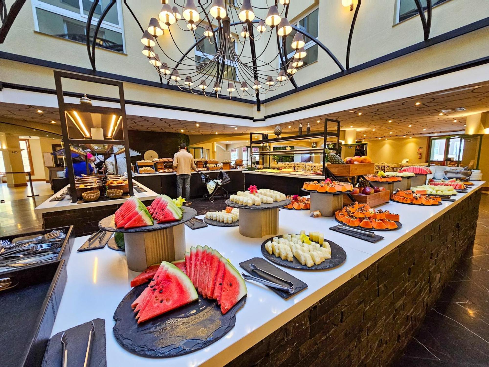 Buona Vitta Gramado Resort & Spa By Gramado Parks Екстер'єр фото The photo depicts a lavish buffet setup in a dining area. In the foreground, there are several platters featuring an assortment of colorful food items, primarily fruits like watermelon and various types of sushi or rolls. The buffet table is elegantl