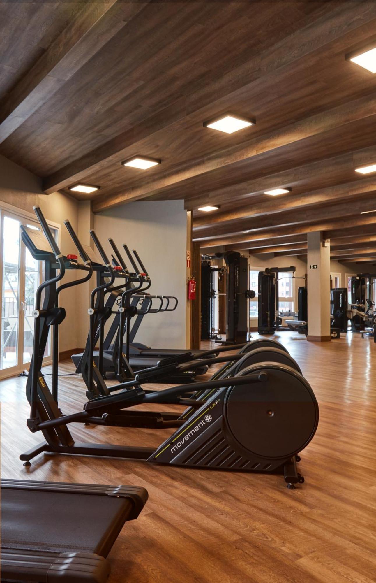 Buona Vitta Gramado Resort & Spa By Gramado Parks Екстер'єр фото The photo shows a modern gym interior with wooden flooring and a ceiling with wooden beams. There are several exercise machines, including elliptical trainers and a treadmill, positioned in the foreground. In the background, there are additional work