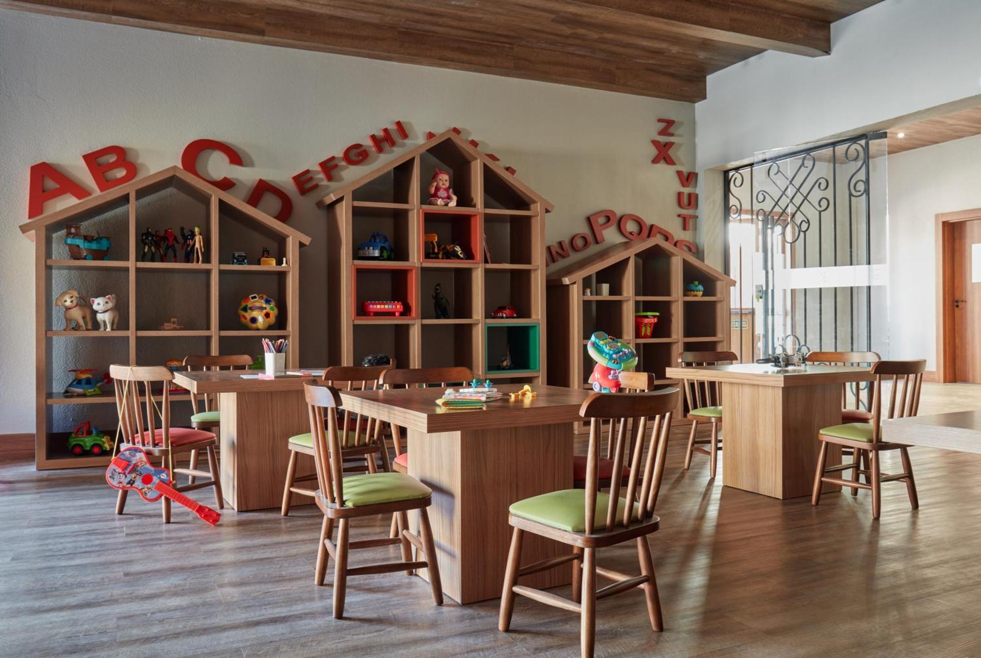 Buona Vitta Gramado Resort & Spa By Gramado Parks Екстер'єр фото The photo shows a classroom or playroom designed for children. It features wooden furniture, including several tables and chairs with green cushions. In the background, there are large, open shelving units shaped like houses, displaying various toys 