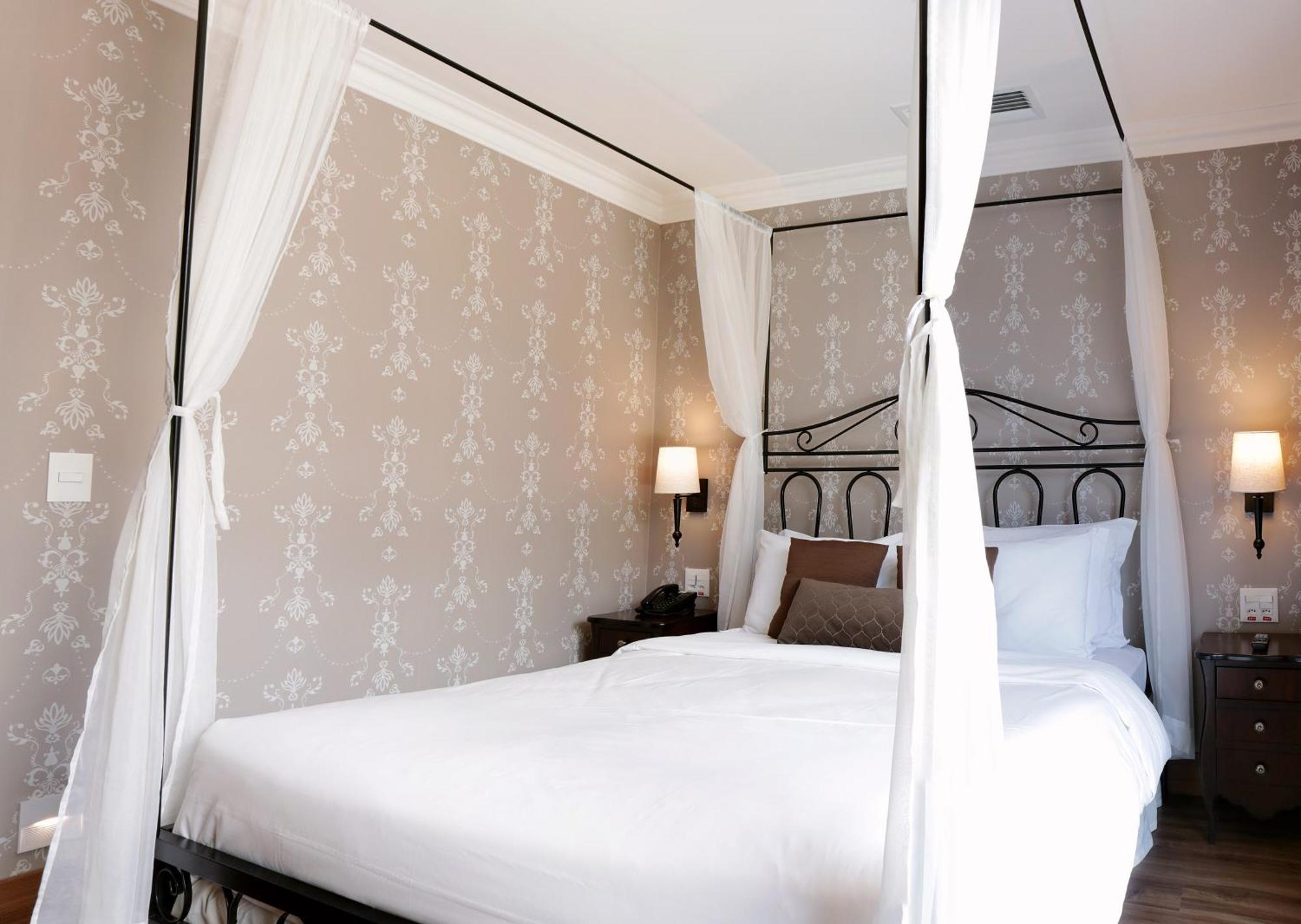 Buona Vitta Gramado Resort & Spa By Gramado Parks Екстер'єр фото The photo shows a elegantly decorated bedroom featuring a four-poster bed with white drapes. The bed is made with white linens and has some brown accent pillows. The walls are adorned with a subtle floral pattern, and there are two bedside lamps prov