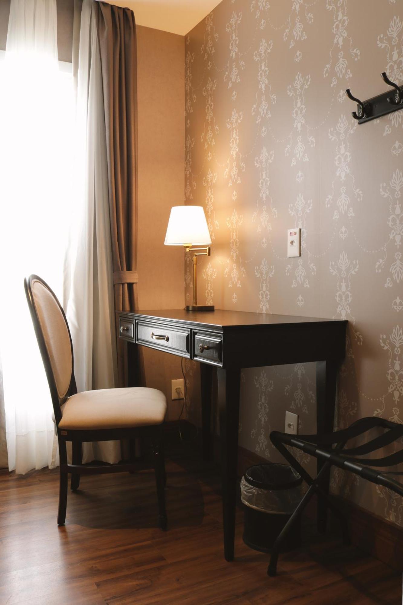 Buona Vitta Gramado Resort & Spa By Gramado Parks Екстер'єр фото The photo shows a small desk set against a wall with a decorative pattern. The desk is black with a few drawers and has a small lamp on it. Next to the desk, there is a chair with a light-colored cushion. Soft light is coming through a window, castin